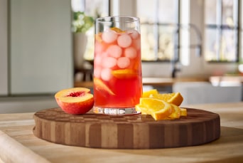 A glass filled with crushed ice and lemon slices is placed on a wooden board, surrounded by whole lemons and green leaves.